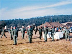 Men in military uniform face each other in rows in a grassy field. There are canvas tents and forest in the background